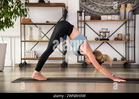 Side view of young flexible female doing Downward Facing Dog asana at home during fitness workout Stock Photo