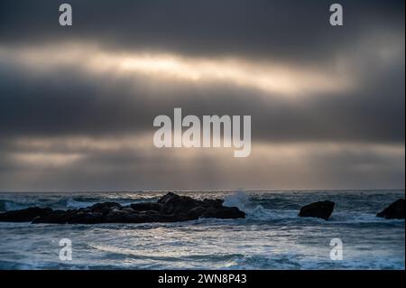 Sun rays coming through gray clouds over ocean waves and coastal rocks Stock Photo