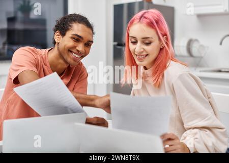 attractive joyous multiracial couple in cozy homewear looking at paperwork while working remotely Stock Photo