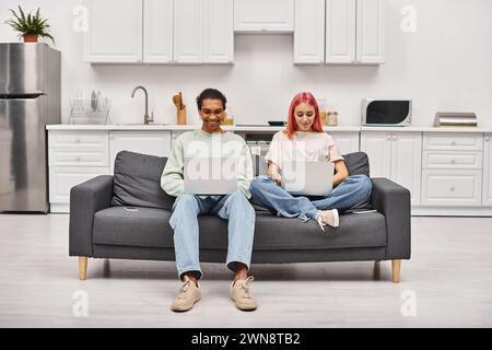 cheerful appealing interracial couple in homewear sitting on sofa and working on laptops remotely Stock Photo