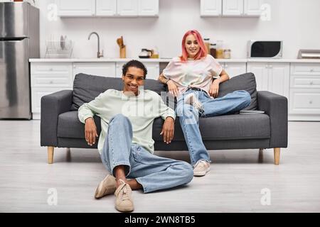 joyous attractive multiracial couple in cozy homewear posing in living room and smiling at camera Stock Photo