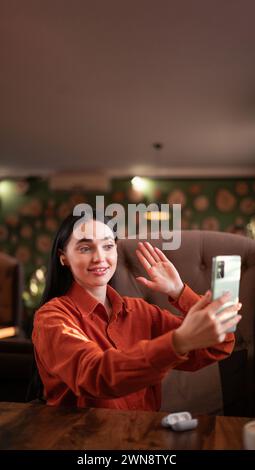 Smiling hipster girl waving hand hello during online video call via cellphone, sitting in cafe. Attractive business woman or student having conference Stock Photo