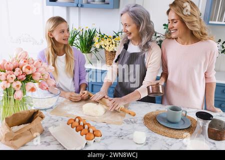 Granddaughter and her mother watching as grandmother makes dough and rolls it out in kitchen table to prepare delicious pastries. Family activity Stock Photo