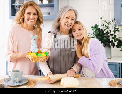 Portrait of women of three generations preparing for Easter holiday. Granddaughter and daughter hugging grandmother who rolls dough for Easter bread Stock Photo