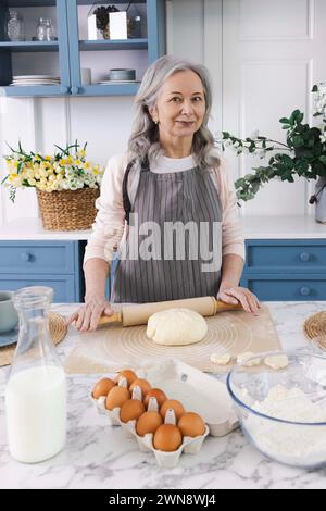 Portrait of elderly woman with gray hair wearing apron in cozy kitchen with rolling pin in her hands making delicious pizza dough. Grandma's dough rec Stock Photo
