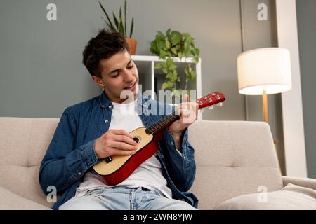 Young relaxed man playing music with ukelele sitting on sofa at home. Young adult carefree male learning to play guitar on couch at the living room. Stock Photo