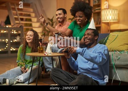 Full length portrait of multiethnic group of adults playing video games emotionally during party at home with African American woman holding controller Stock Photo