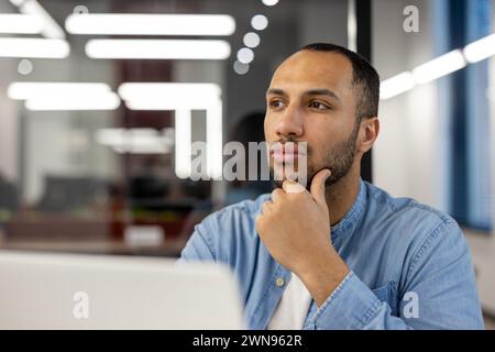 A contemplative young professional in a well-lit office environment, caught in a moment of thoughtfulness while gazing away from his laptop screen. His expression conveys concentration and deep thinking. Stock Photo