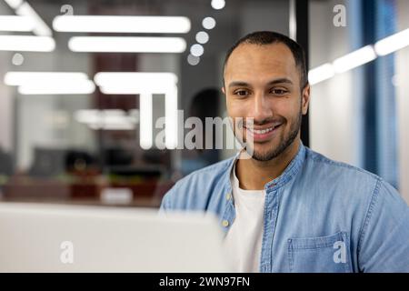 A young, professional man wearing a denim shirt smiles confidently at the camera from his workstation in a contemporary office environment, illuminated by natural and artificial light, embodying ambition and approachability. Stock Photo