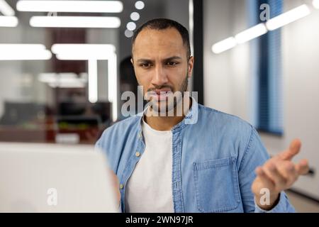 A young male professional in a blue denim shirt appears perplexed and frustrated while working at his computer in a modern office setting, expressing a common workplace challenge. Stock Photo
