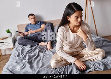 frustrated asian woman sitting near husband using smartphone in bedroom at home, family problem Stock Photo
