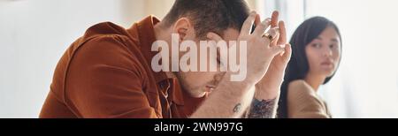 young tattooed sad man sitting with bowed head near young asian woman, divorce concept, banner Stock Photo