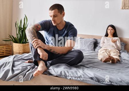 upset tattooed man sitting on bed near frustrated asian wife, family relationship difficulties Stock Photo