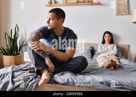 frustrated tattooed man sitting on bed near upset asian wife, family relationship difficulties Stock Photo