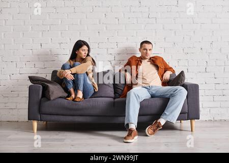 frustrated asian woman sitting near self-assured husband on couch in living room, misunderstanding Stock Photo