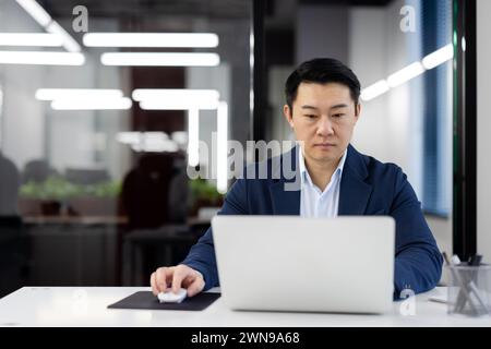 Busy formal office worker sitting by desktop with modern laptop and using wireless digital mouse on blurred background. Korean manager checking mailbox of marketing agency and clicking on refresh tab. Stock Photo