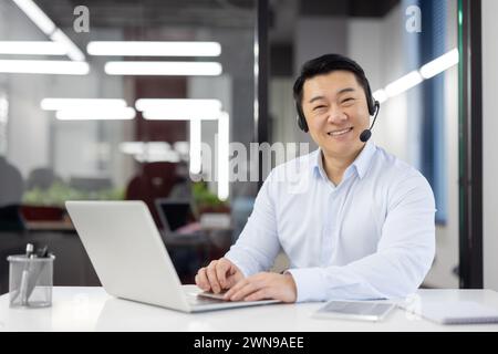 Positive office clerk in white shirt and black headset sitting by desk with portable computer at workspace. Carefree chinese person spending busy day in coworking with happy facial expression. Stock Photo