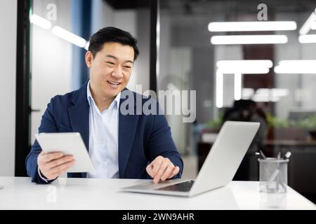 Modern businessman in formal clothes using digital tablet while working by pc on white table. Successful entrepreneur using two wireless devices for comparing articles from different web sources. Stock Photo
