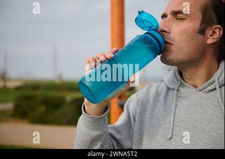 Close-up sportswoman, male athlete drinks water to hydrate body after outdoor cardio workout. Middle aged athletic man resting after jogging at dawn. Stock Photo