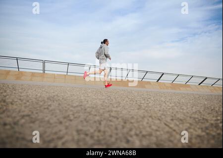 Side view of a determined muscular build young sportsman, male athlete jogger in gray active wear and red sports shoes, running on the city treadmill, Stock Photo