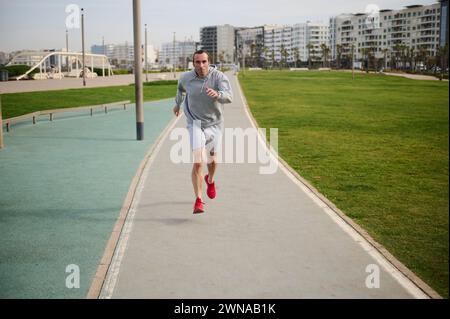 Full length shot of healthy young man in gray sportswear and red sneakers, running on the promenade. Male runner sprinting on the treadmill outdoors. Stock Photo