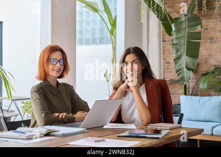 Portrait of two happy business women sitting at work desk with laptop. Stock Photo