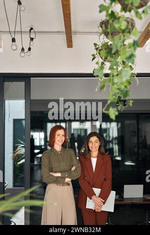 Two happy smiling business women standing at work in office. Vertical portrait. Stock Photo