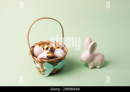 Basket with painted white and golden eggs and bunny on light green background. Easter concept. Closeup. Stock Photo