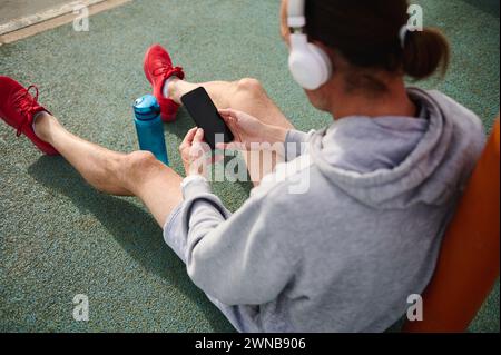 Overhead view of active sports man wearing wireless headphones, checking sports app on his smart mobile phone, sitting on the sportsground. Cell phone Stock Photo