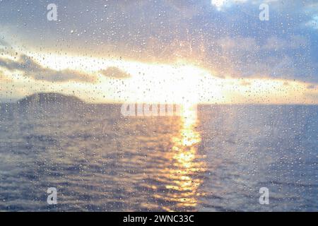 View of a sea sunset through raindrops on a window pane. Raindrops on window pane on sea sunset background. Stock Photo