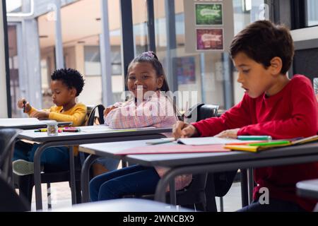 Three children are focused on coloring at desks in a school classroom setting Stock Photo