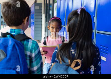 Biracial girl with pink headband talks to her peers by blue school lockers, holding a notebook Stock Photo