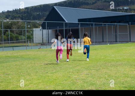 Three young girls are running across a grassy field near a building with copy space in school Stock Photo