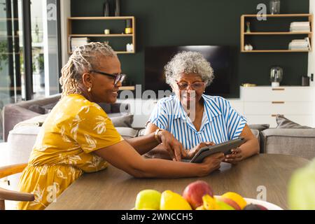 Two senior women share a tablet in a cozy, decorated living room. Stock Photo