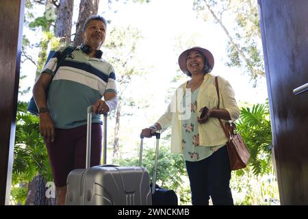 Senior biracial couple in casual travel attire ready for a trip, with luggage in hand Stock Photo