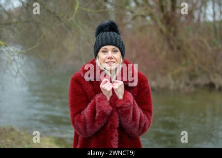 Attractive blonde lady with red jacket and black bobble hat holds her coat with her hands because it is so cold Stock Photo