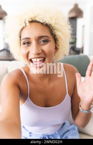 Young biracial woman with curly blonde hair smiles joyfully, waving at the camera on a video call Stock Photo