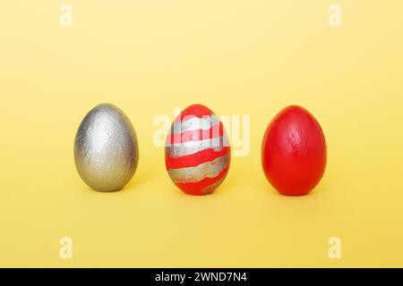 abstract composition of three multi-colored handmade eggs on a yellow pastel background. Eggs of metallic, red, red with gray stripes on a yellow pape Stock Photo