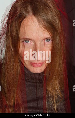 Vertical composition. Portrait of an adult young woman with long blond hair and gray eyes and a red transparent cloth on her head. Stock Photo