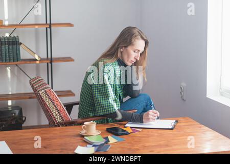 A young pretty lady is thinking and taking notes on a tablet against the backdrop of a work environment on the table. Stock Photo