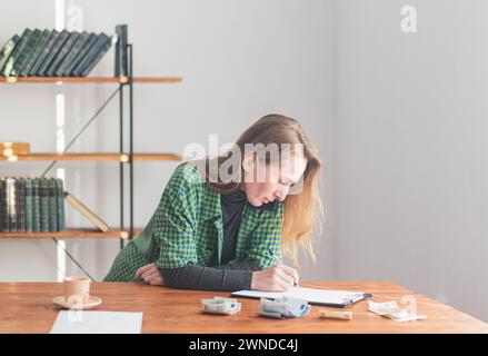 A young pretty lady speaks on the phone and writes on a tablet against the backdrop of a work environment and money laid out on the table. Stock Photo