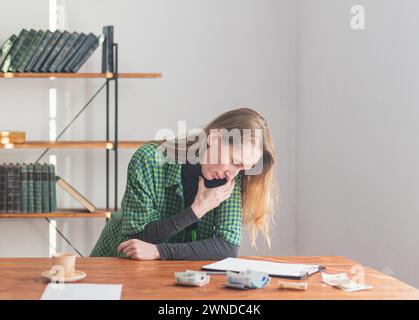 A young pretty lady is talking on the phone and looking at notes on a tablet against the backdrop of a work environment and money laid out on the tabl Stock Photo