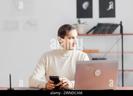 A young businessman in a bright office is thoughtful and looks intently to the side while holding a smartphone. Stock Photo