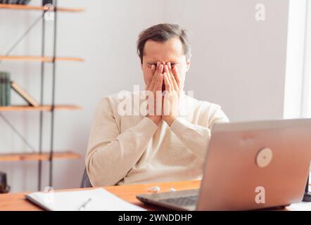 A young adult man in a home office working on a laptop is tired and covers his eyes with his hands. Stock Photo