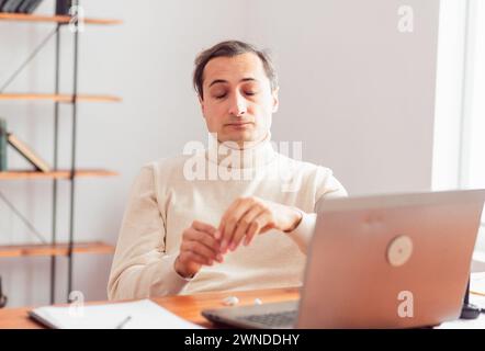 A young adult man in a home office is thoughtful and closed his eyes while working on a laptop and mobile phone. There are graphic electronic and pape Stock Photo