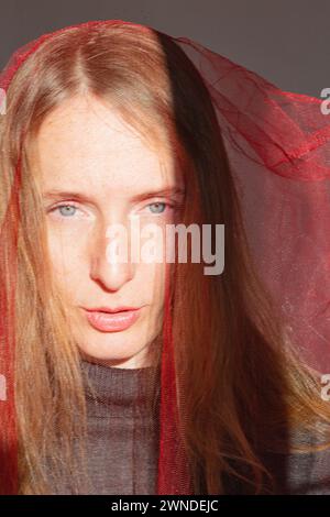 Vertical composition. Portrait of an adult young woman with long blond hair and gray eyes in the bright rays of the sun and a red transparent cloth on Stock Photo