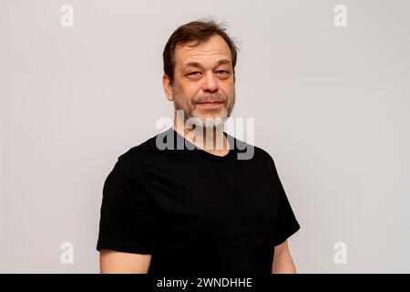 An elderly man 50-55 years old with a beard on a white background, wearing a black T-shirt, looks confidently into the camera. Stock Photo