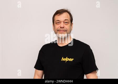 Elderly man 50-55 years old wearing a black T-shirt that says: Helper. Looking into the camera on a white background. Stock Photo