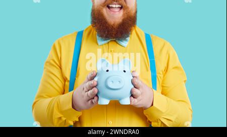 Piggy bank in form of pig in hands of joyful stylish fat man on light blue background. Stock Photo