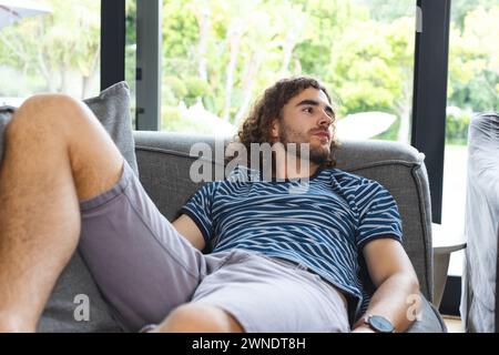 Young Caucasian man with curly hair lounges on a gray sofa, gazing thoughtfully Stock Photo